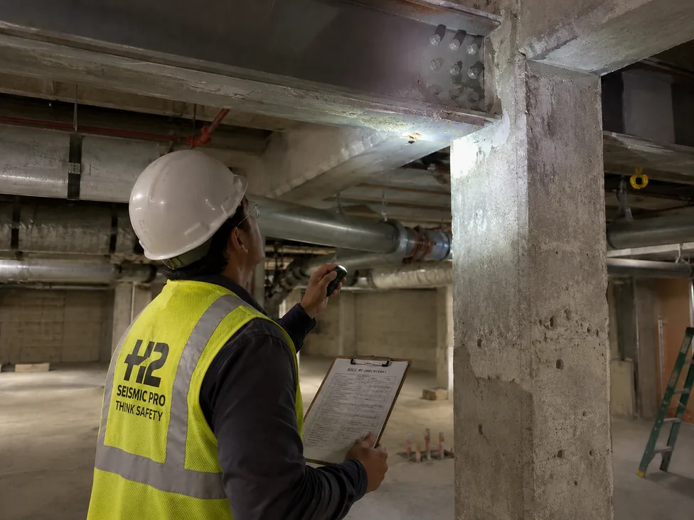 H2 Seismic Pro engineer in a hard hat and high-vis vest inspecting an exposed steel beam-to-concrete-column connection in a building basement during an ASCE 41-23 tiered seismic evaluation, holding an ASCE 41 checklist clipboard.