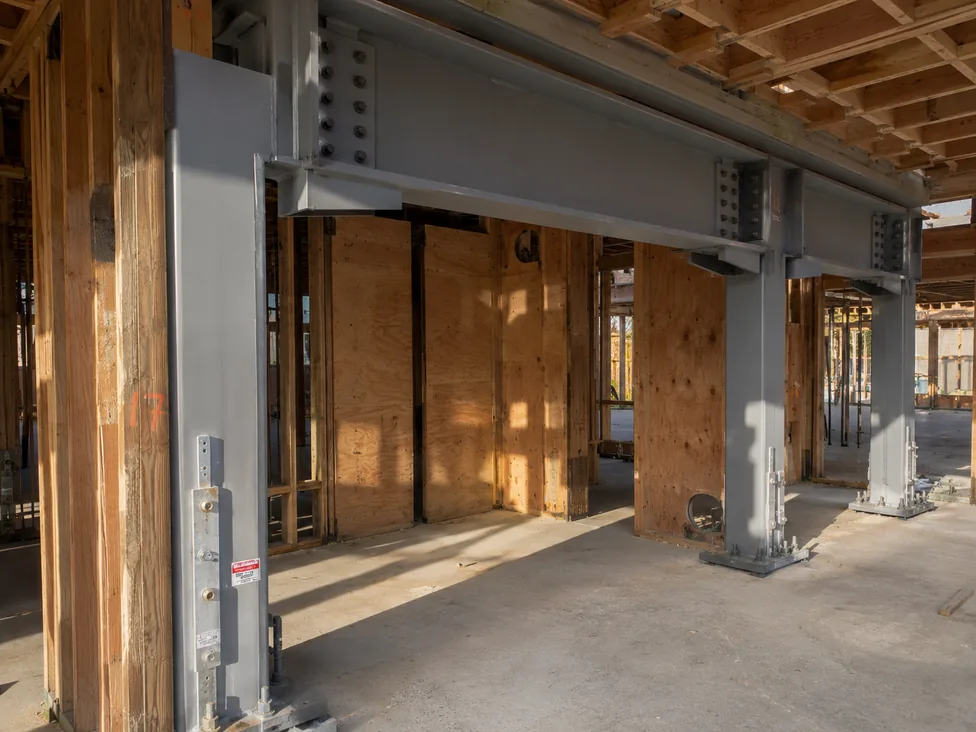 Steel moment frame retrofit installed in a soft-story wood-frame multifamily ground floor — painted gray HSS columns and W-section beam with bolted gusset connections and hold-downs anchored to new concrete pads. PE-stamped per ASCE 41-23.