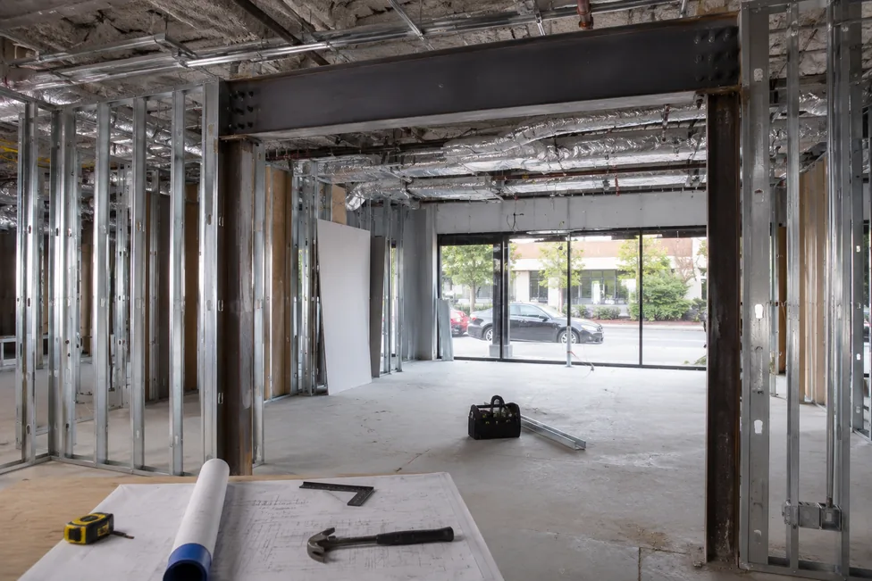 Commercial tenant improvement build-out in progress — new metal-stud framing for partitions with a structural steel header beam spanning a widened storefront opening, rolled drawings and tools on a workbench in the foreground.